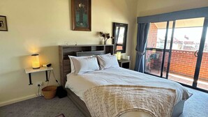 Un dormitorio con cama, lámpara de noche, un cuadro enmarcado y vistas por la ventana.