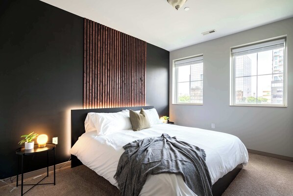 A modern bedroom with a large bed, a bedside table with a lamp, and a view of the city through the windows.