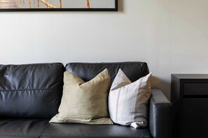 A black leather sofa with two pillows, one beige and one grey, against a plain wall with a framed picture above.