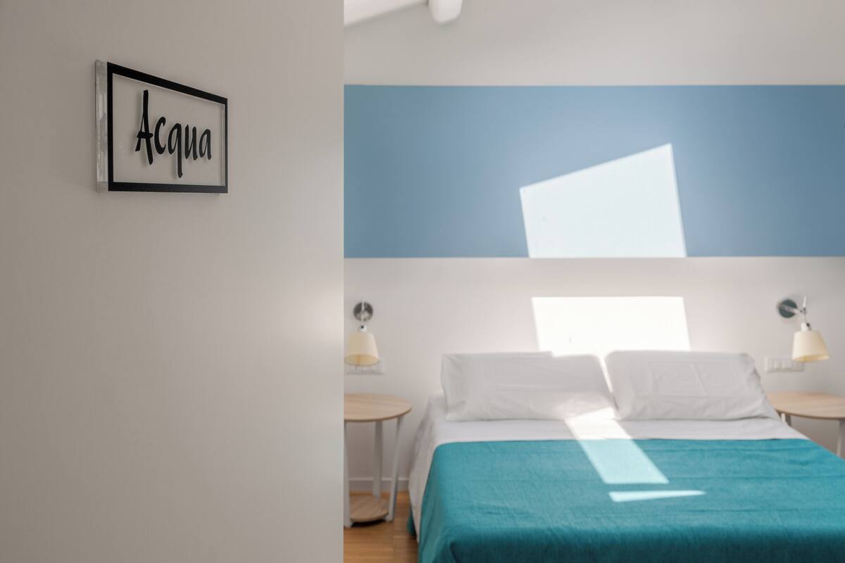 A bedroom with a bed, two bedside tables, a wall-mounted lamp, and a framed sign on the wall.