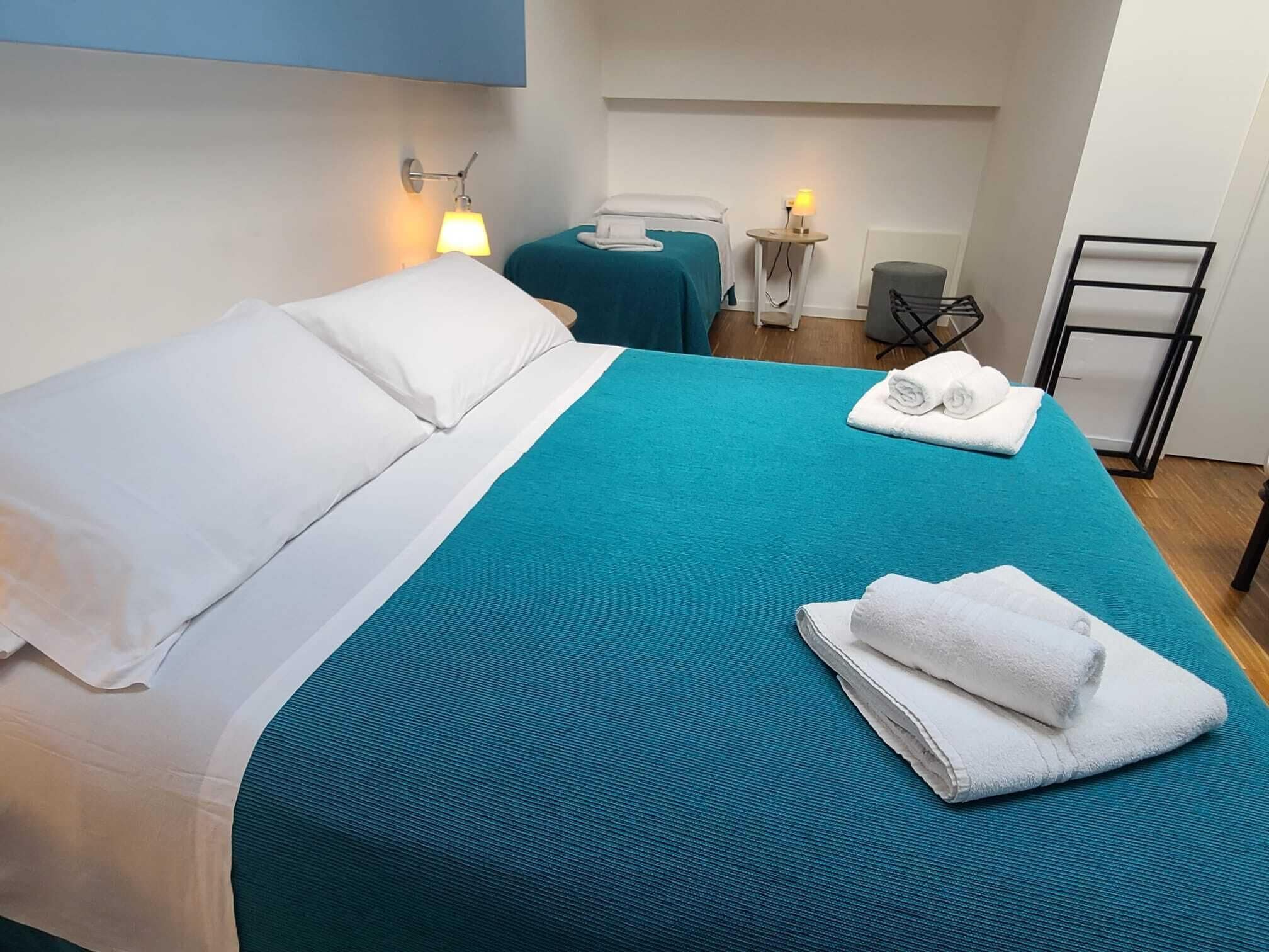 Hotellihuone, jossa on kaksi sänkyä, pieni pöytä, tuoli ja pyyheteline.