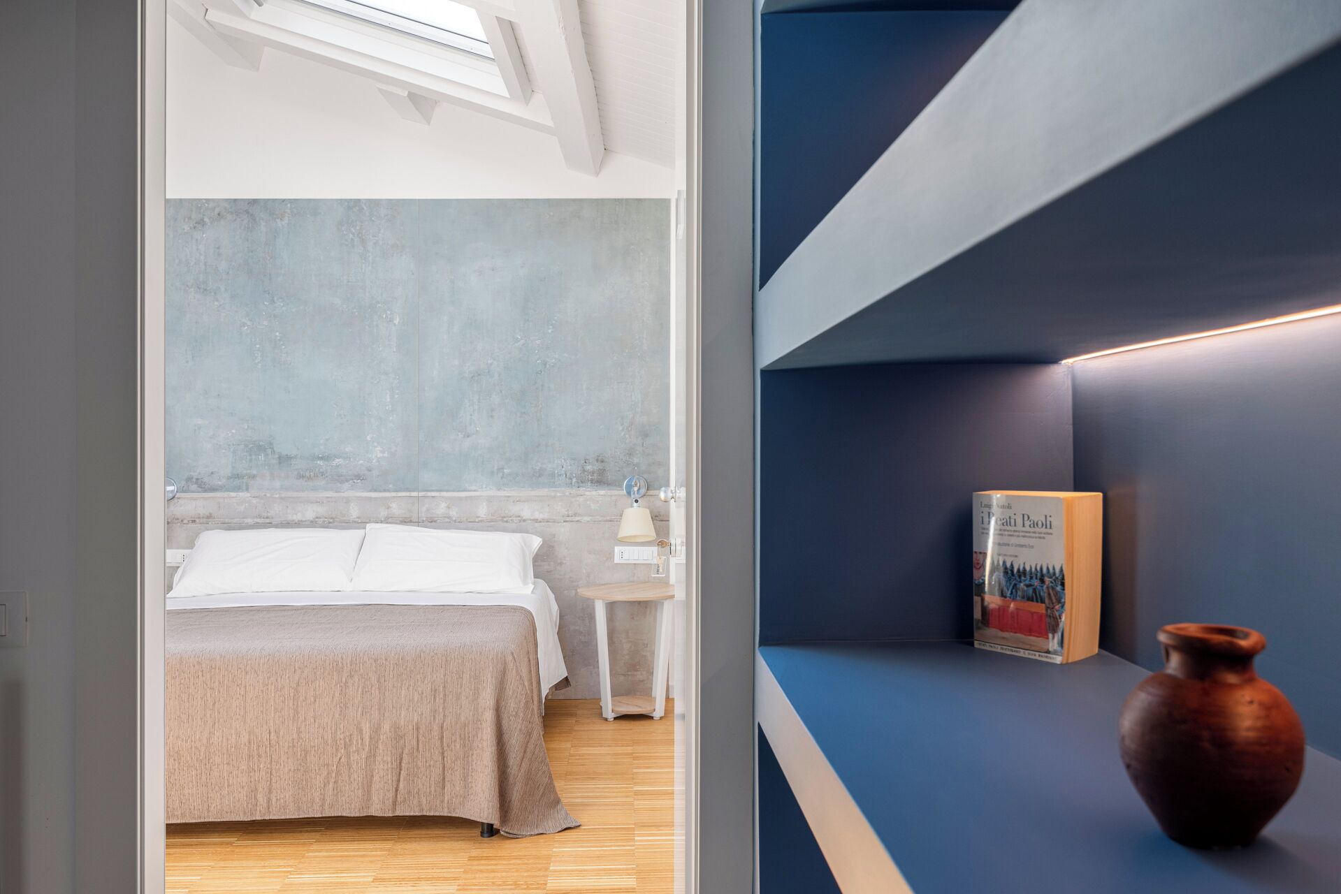 Um quarto moderno com uma cama, mesa de cabeceira e criado-mudo. Há um livro e um vaso em uma prateleira.