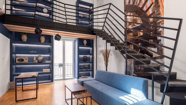 A modern living room with a blue sofa, wooden flooring, a staircase, and a decorative wall with shelves. - Le Stanze di Triyah (Palermo)