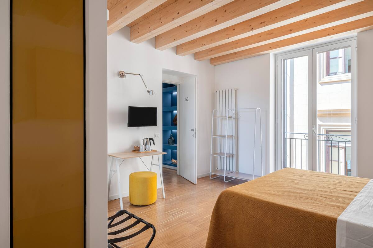 A modern bedroom with a wooden ceiling, a bed with a brown bedspread, a desk with a computer, a yellow ottoman, and a view of the outside through a large window.