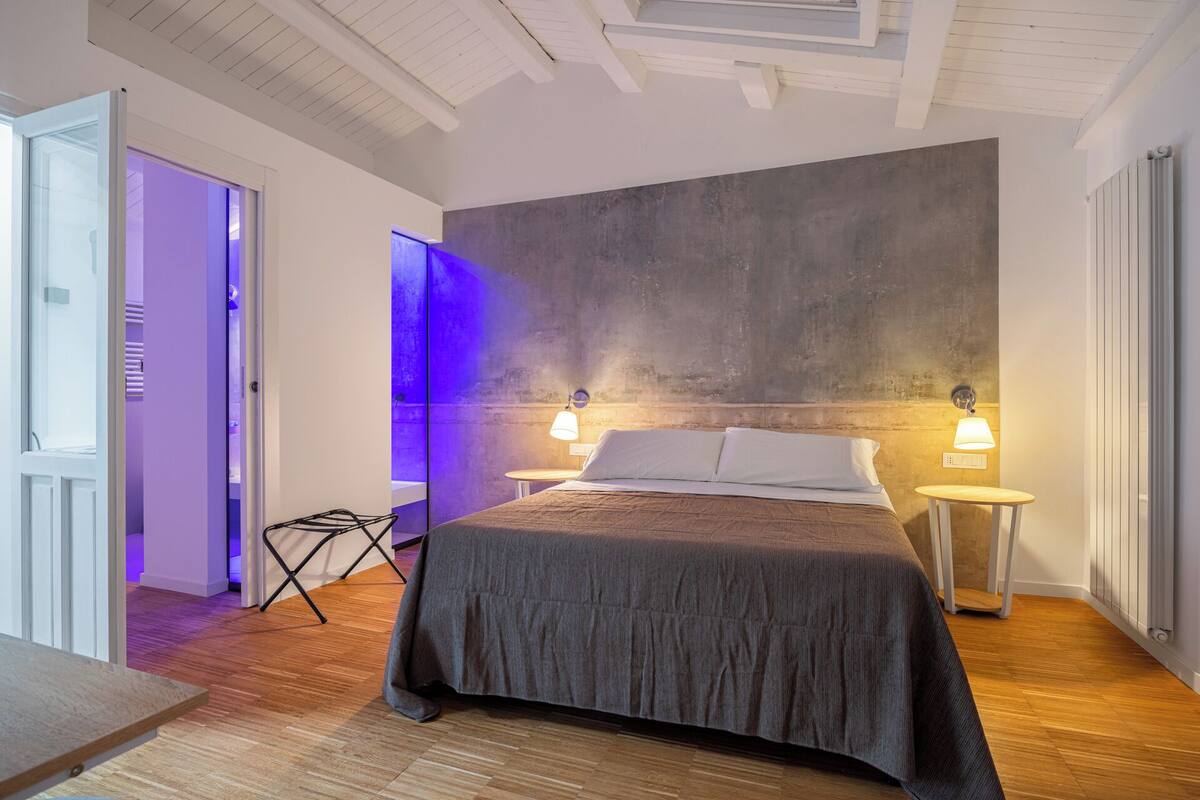 A modern bedroom with a large bed, two bedside lamps, and a view of a bathroom with purple lighting.