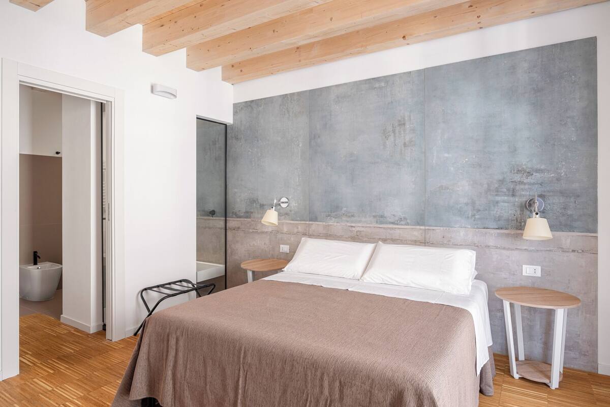 A modern bedroom with a large bed, bedside tables, a mirror, and a concrete accent wall.