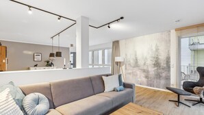 A modern living room with a sofa, coffee table, and a chair. There is a kitchen area in the background with a counter and cabinets. - GebirgsResort Winterberg (Winterberg)