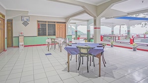 A covered outdoor dining area with tables and chairs, a tiled floor, and a view of a red and green structure in the background. - Hotel O Lie Mas Hotel (Prigen)