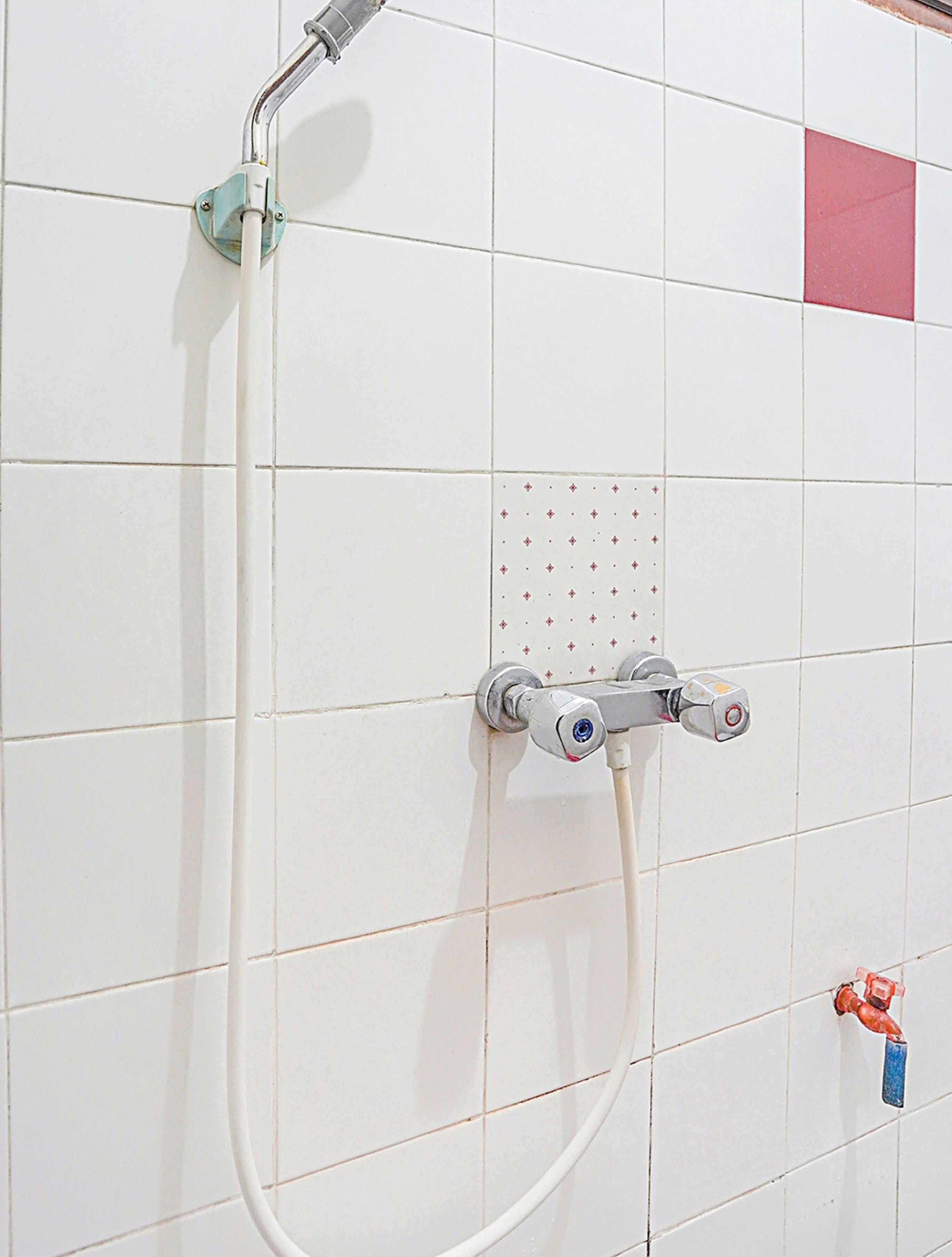 Un baño con paredes de azulejos blancos, un cabezal de ducha, una ducha de mano y un grifo con asa roja.