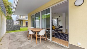 Une terrasse extérieure moderne avec une table en bois et des chaises blanches, un mur en pierre et une vue sur l’intérieur.