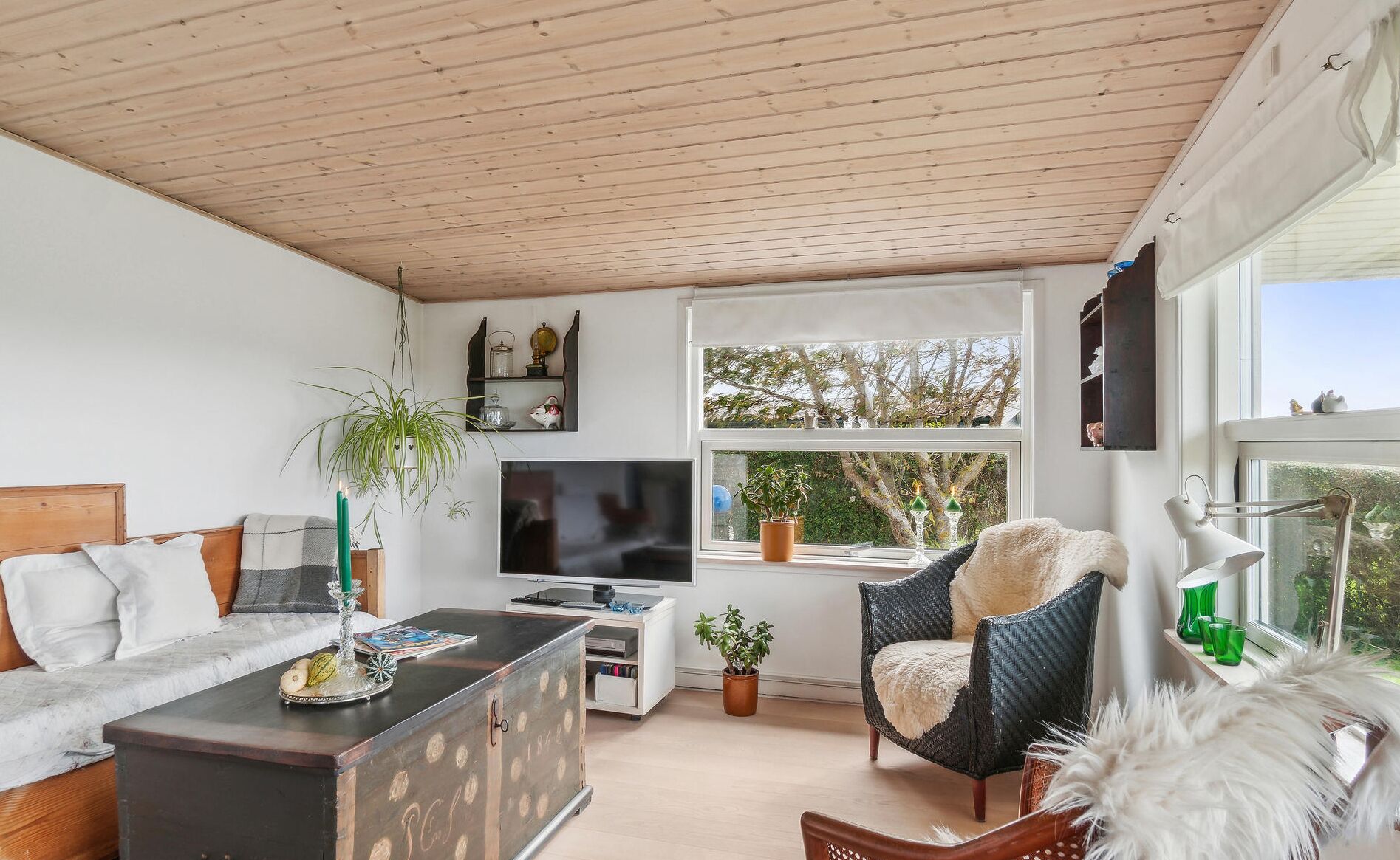 Una sala de estar acogedora con techo de madera, un sofá, un televisor, una estantería y un ventanal con vistas a árboles.