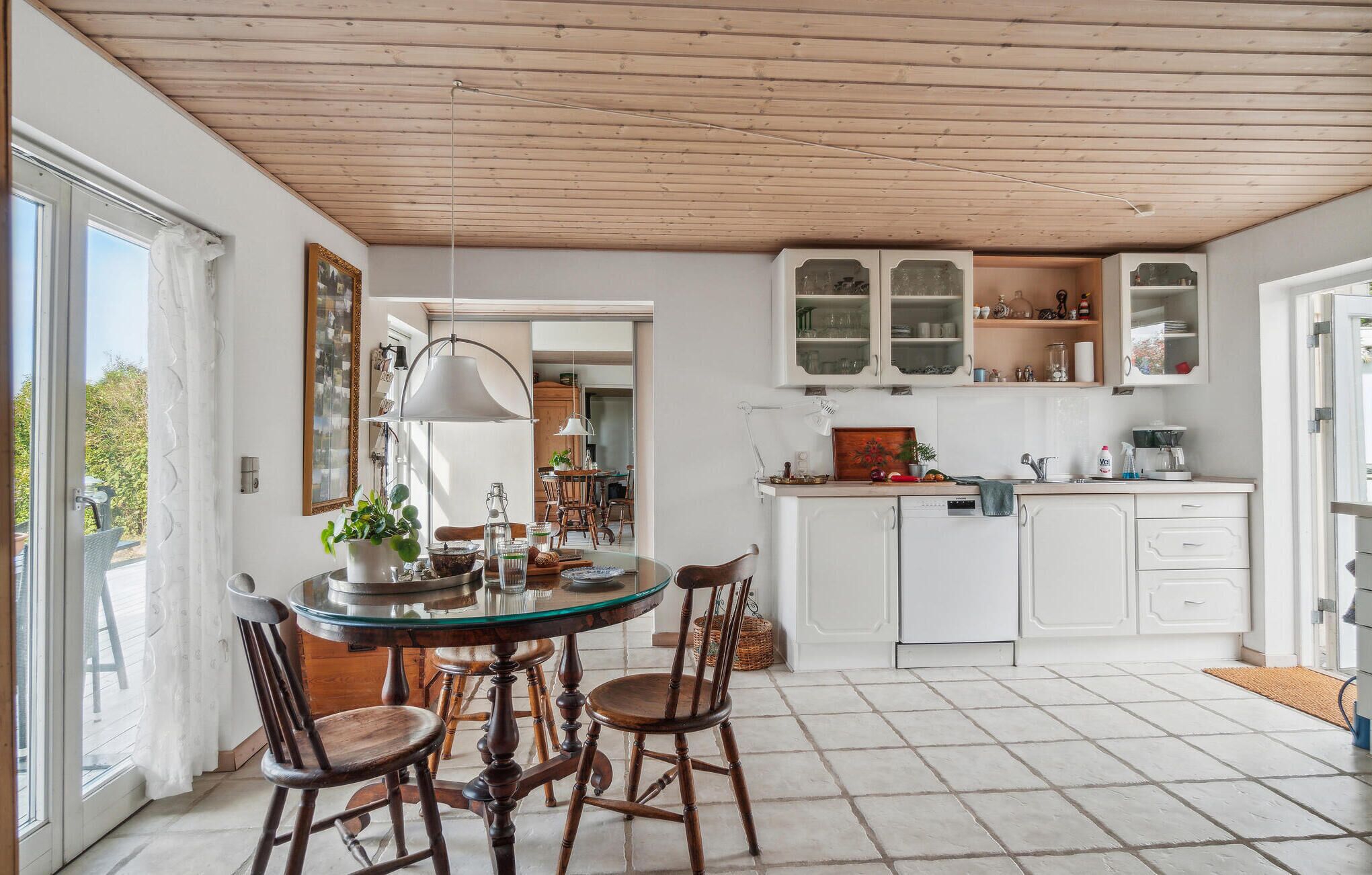 Una cocina con una mesa redonda de vidrio, sillas de madera, armarios blancos y techo de madera.