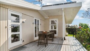 Katettu terassi, jossa on puinen lattiapinta, pöytä ja tuolit sekä lasipaneeleilla varustettu ovi.