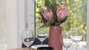 Una mesa de comedor dispuesta con platos blancos, copas de vino y un jarrón rosa con flores.