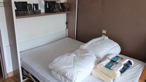 Un lit bien fait, avec une literie et des oreillers blancs, une petite table de chevet sur laquelle se trouvent un livre et une boîte de mouchoirs, et un tableau accroché au mur.