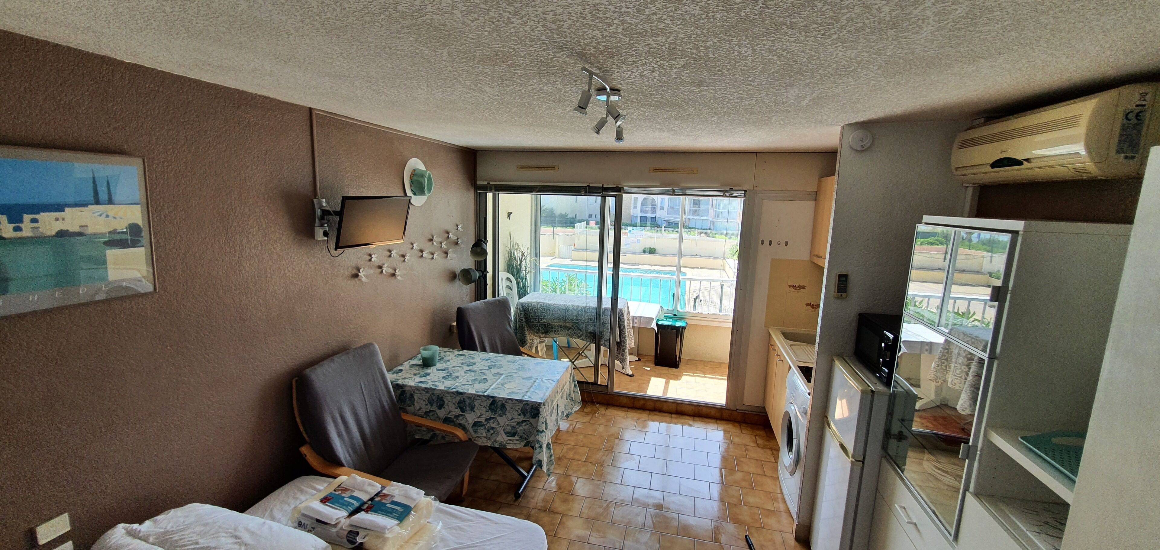 A compact living space with a kitchenette, a dining area with a table and chairs, a small sofa, a ceiling fan, and a view of a swimming pool through a glass door.