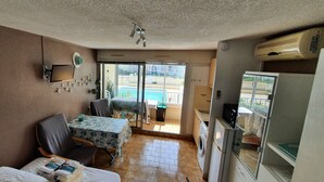 Un espace de vie compact comprenant un coin cuisine, un espace repas avec une table et des chaises, un petit canapé, un ventilateur de plafond et une vue sur une piscine, visible à travers une porte vitrée.