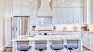 A kitchen with white cabinets, a stainless steel refrigerator, a marble island with bar stools, and pendant lighting. - Gulfport's Premier Beachfront Getaway, Ocean View Beach House! Sleeps 10 (Gulfport)