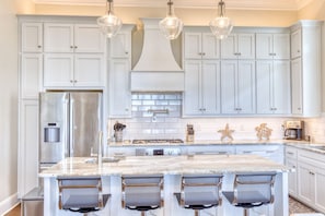 Une cuisine moderne avec des meubles blancs, un réfrigérateur en acier inoxydable, un îlot en marbre avec des tabourets de bar et des luminaires suspendus.