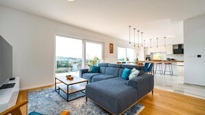 A modern living room with a grey sectional sofa, a small coffee table, and a dining area with a bar. The kitchen is visible with white cabinets and a large window. - Stunning home in Zadar, Pasman (Zadar, Pasman)