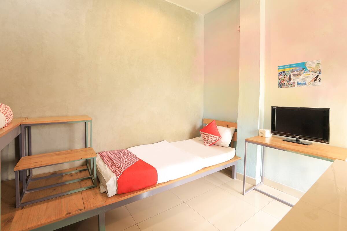 A single bed with a red and white blanket, a wooden bedside table, a TV on a stand, and a poster on the wall.