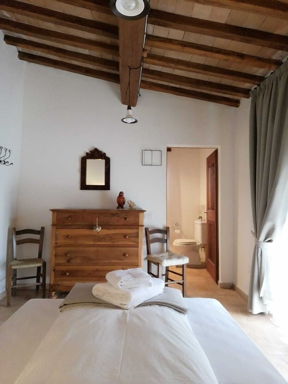 Een slaapkamer met een houten dressoir, twee stoelen, een bed met netjes gevouwen handdoeken, een spiegel en een plafondlamp.