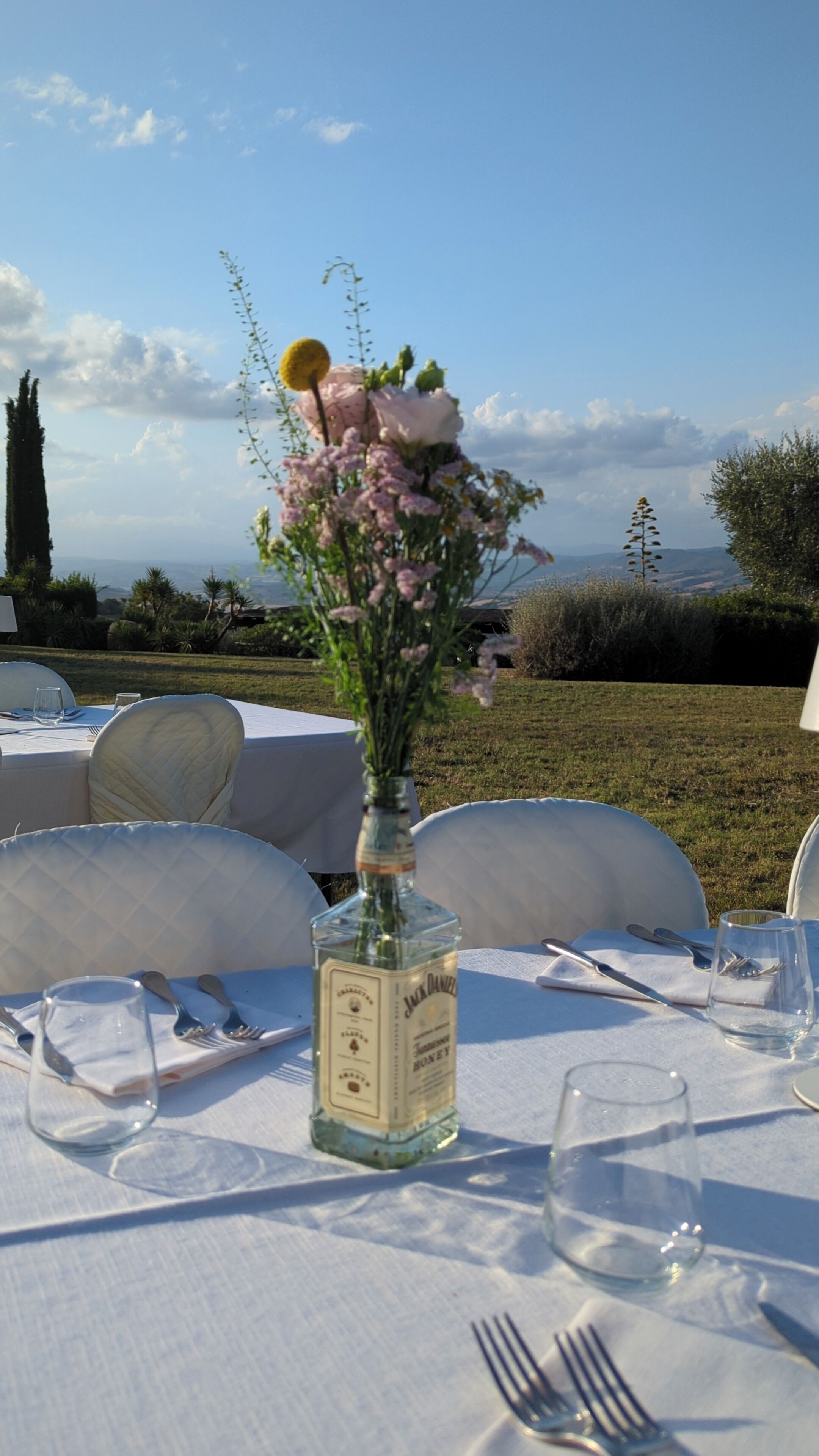 A table set outdoors with a bottle of Jack Daniel's whiskey, a vase with flowers, and a clear view of the surrounding landscape.