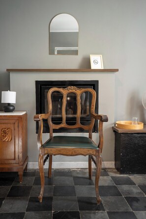 A wooden chair with a green cushion, a fireplace with a mirror above, and a small wooden table with a lamp.