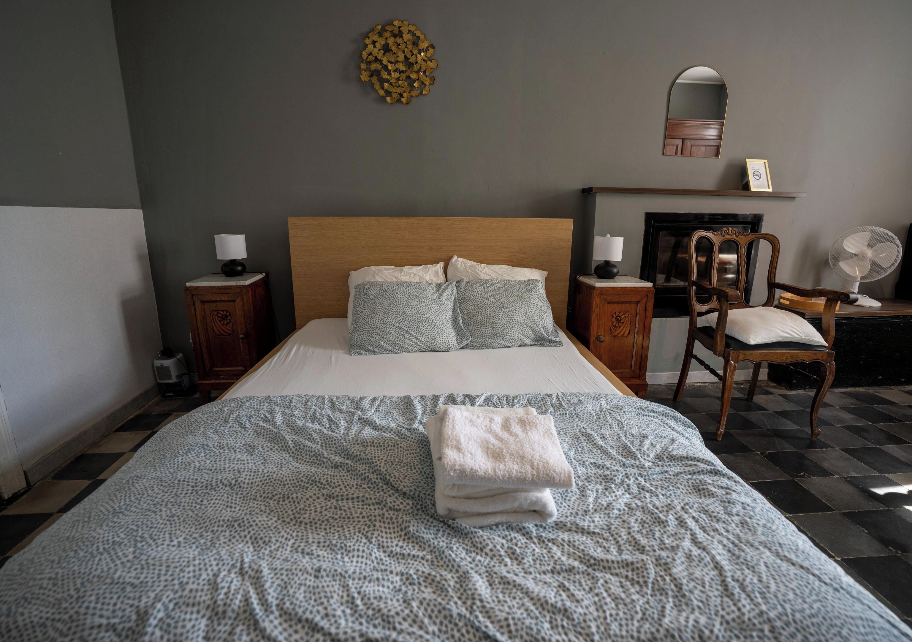 En pænt redt seng med et mønstret sengetæppe og puder, et natbord med en lampe, en stol, et spejl og en vægmonteret hylde med en ramme.