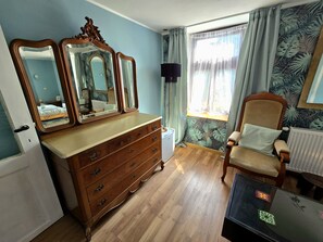 Ein Schlafzimmer mit einem Holzkoffer, einem Stuhl, einem Spiegel und einem Fenster mit Vorhängen.