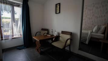 A room with a dining table, two chairs, a bed with patterned bedding, a window with sheer curtains, and a wall-mounted picture frame.