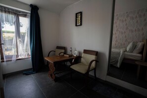 A room with a dining table, two chairs, a bed with patterned bedding, a window with sheer curtains, and a wall-mounted picture frame.