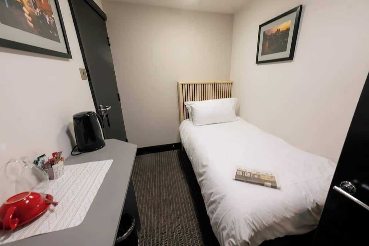 A hotel room with a bed, a desk with a kettle, a red cup, and a framed picture on the wall.