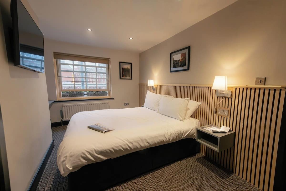 A hotel room with a large bed, a bedside table with a lamp, a window with blinds, and a radiator.