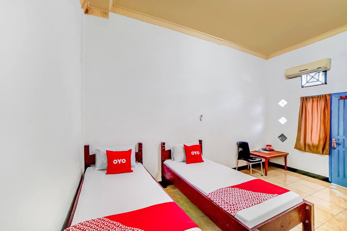 Une chambre d’hôtel avec deux lits simples, chacun recouvert d’une couvre-lit rouge et blanche, assortie aux oreillers. On y trouve un bureau avec une chaise et une petite table sur laquelle se trouve un objet rouge. Un climatiseur est fixé au mur, et une fenêtre avec des rideaux est visible.
