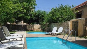 Une piscine rectangulaire à l’eau d’un bleu limpide, entourée d’une terrasse en béton. On y trouve deux chaises longues blanches à armature métallique et un escalier en métal qui mène à la piscine. La zone est ombragée par des arbres et un mur en béton se dresse à l’arrière-plan.