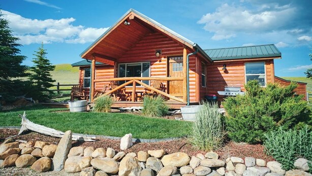 A log cabin with a porch, a grill, and a stone wall in the foreground.