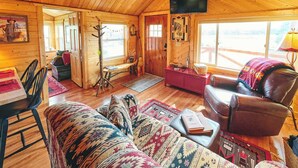 A cozy log cabin interior with a wooden dining table, leather sofa, and a television mounted on the wall.