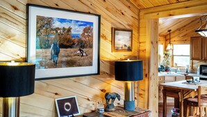 Un intérieur en bois agrémenté d’une photographie de paysage encadrée, d’une lampe de table, d’une petite table avec un ordinateur portable, d’une statuette de cheval décorative et d’un coin cuisine visible à travers une porte ouverte.