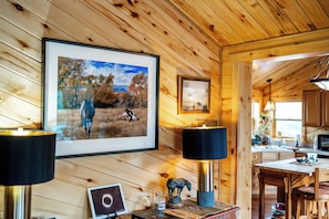 Interior em madeira com uma fotografia emoldurada de paisagem, um abajur, uma pequena mesa com um laptop, uma estatueta decorativa de cavalo e uma cozinha visível através de uma porta aberta.