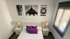 A neatly made bed with white bedding and a rolled towel, two bedside tables with lamps, a window with blinds, and three wall-mounted pictures including the Eiffel Tower. - Reyna 2 Dormitorios (Málaga)
