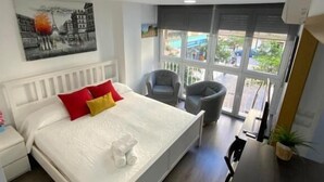 Un dormitorio moderno con cama blanca, almohadas rojas y amarillas, un cuadro de un paisaje urbano, un balcón con sillas y una mesa de comedor con sillas.