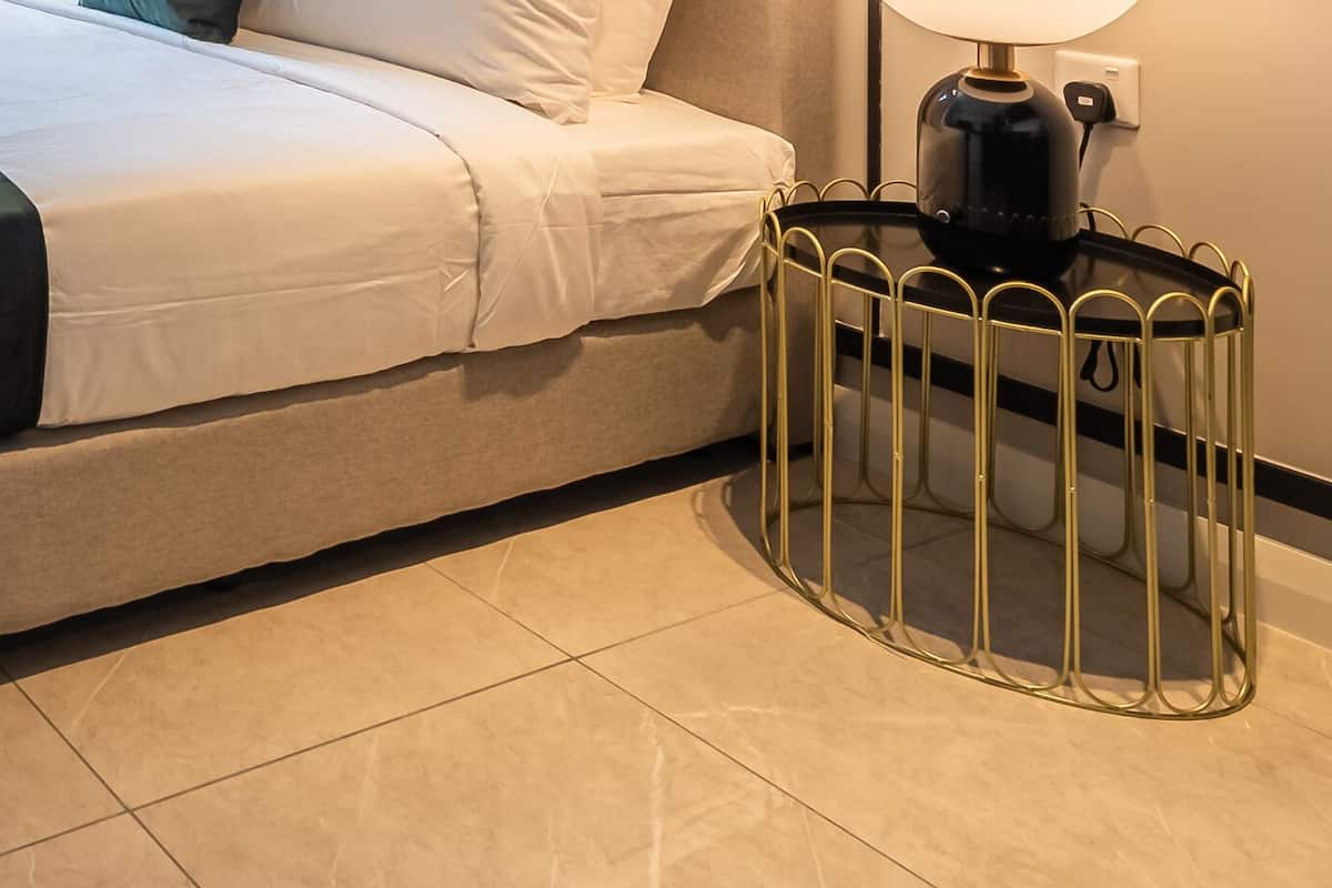 Una cama bien tendida con una manta oscura, una mesita de noche con una lámpara y un cubo de basura con una verja metálica decorativa.