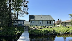 Una casa con un muelle que se adentra en un lago tranquilo, rodeada de exuberante vegetación y un cielo azul despejado.