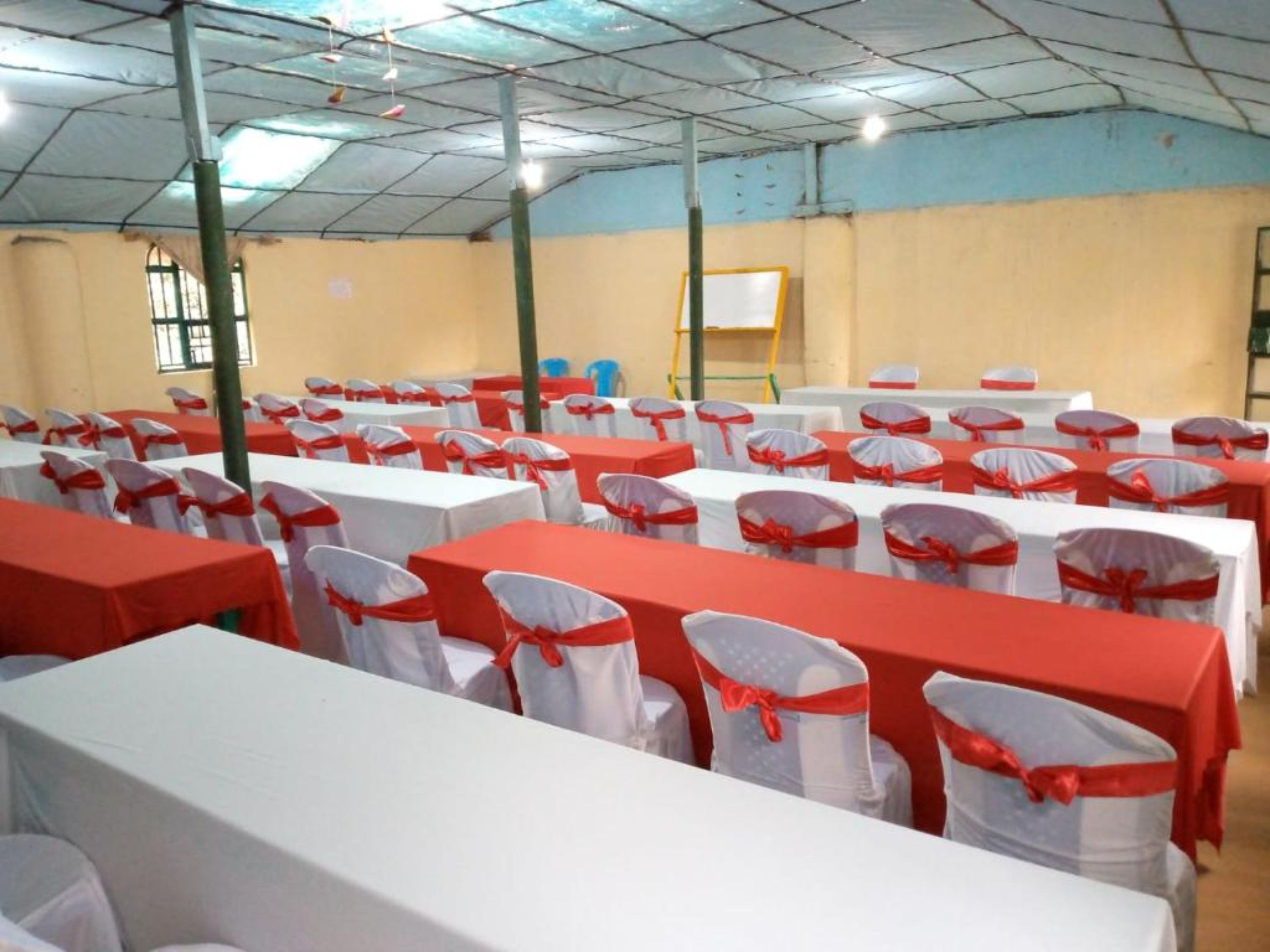 Een congreszaal met rijen tafels, bedekt met witte tafelkleden en rode linten, stoelen met witte hoezen en rode linten, en een whiteboard aan de muur.