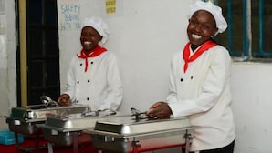 Two individuals in white chef uniforms with red scarves standing in a kitchen area with stainless steel serving trays and a red floor. - Sofa Camp Naivasha (Naivasha)