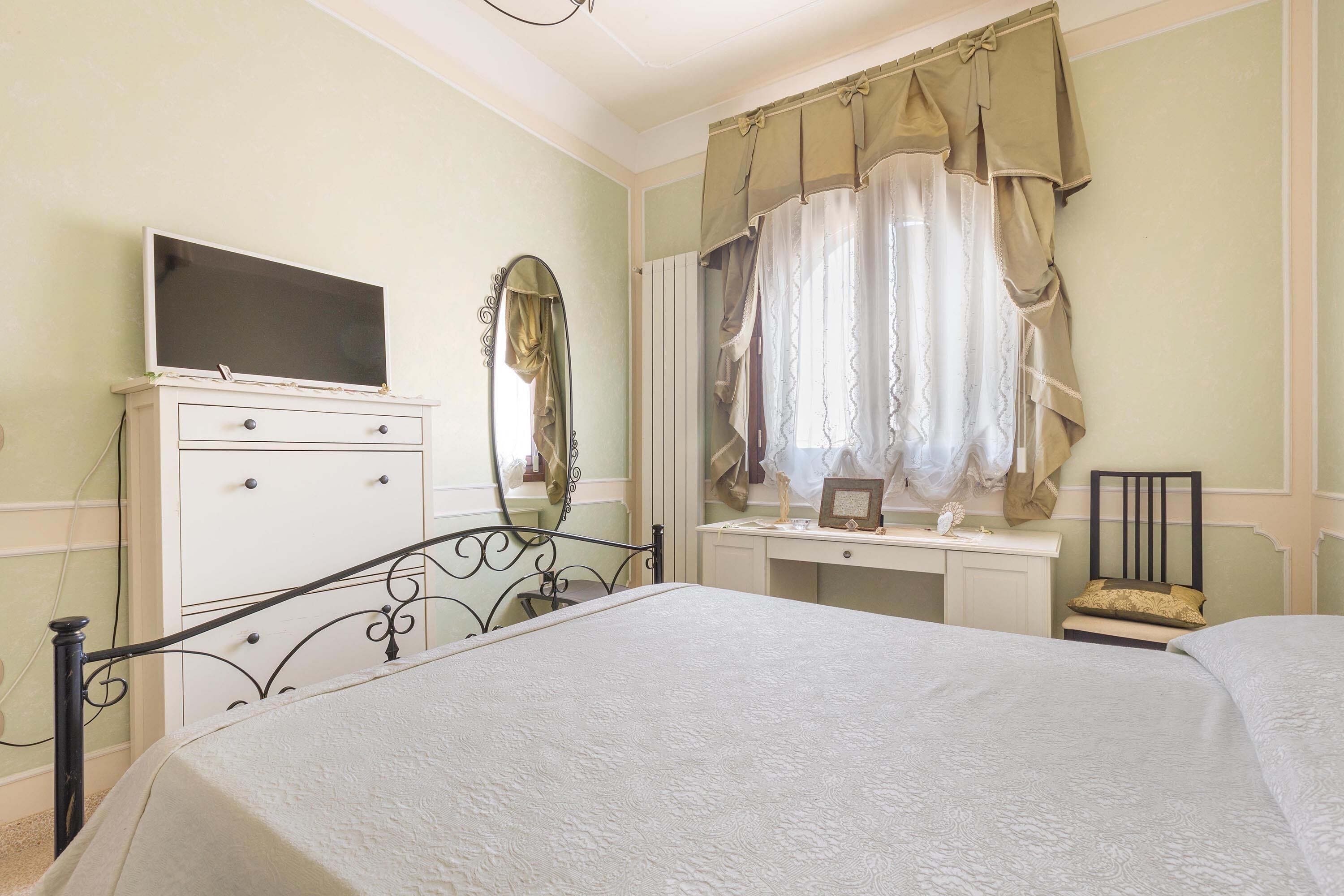 Quarto com cama, cômoda, penteadeira com espelho e televisão sobre a cômoda.