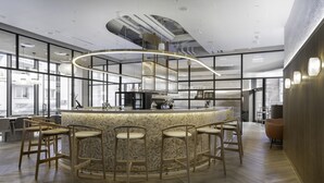Un área de bar moderna con una isla central y taburetes, ventanas amplias y un techo alto con conductos expuestos.