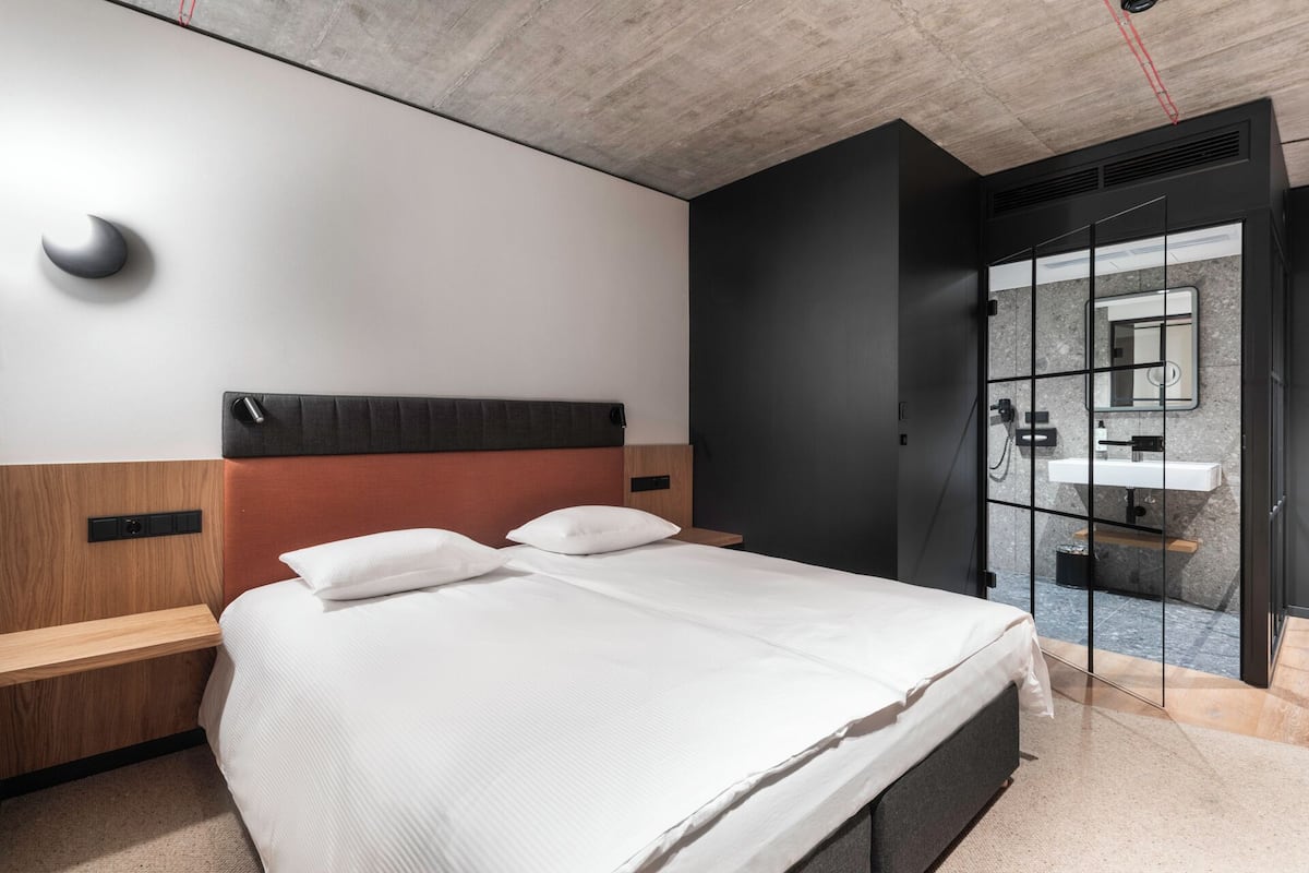 A modern hotel room with a large bed, wooden headboard, and a view of the bathroom through a glass door.