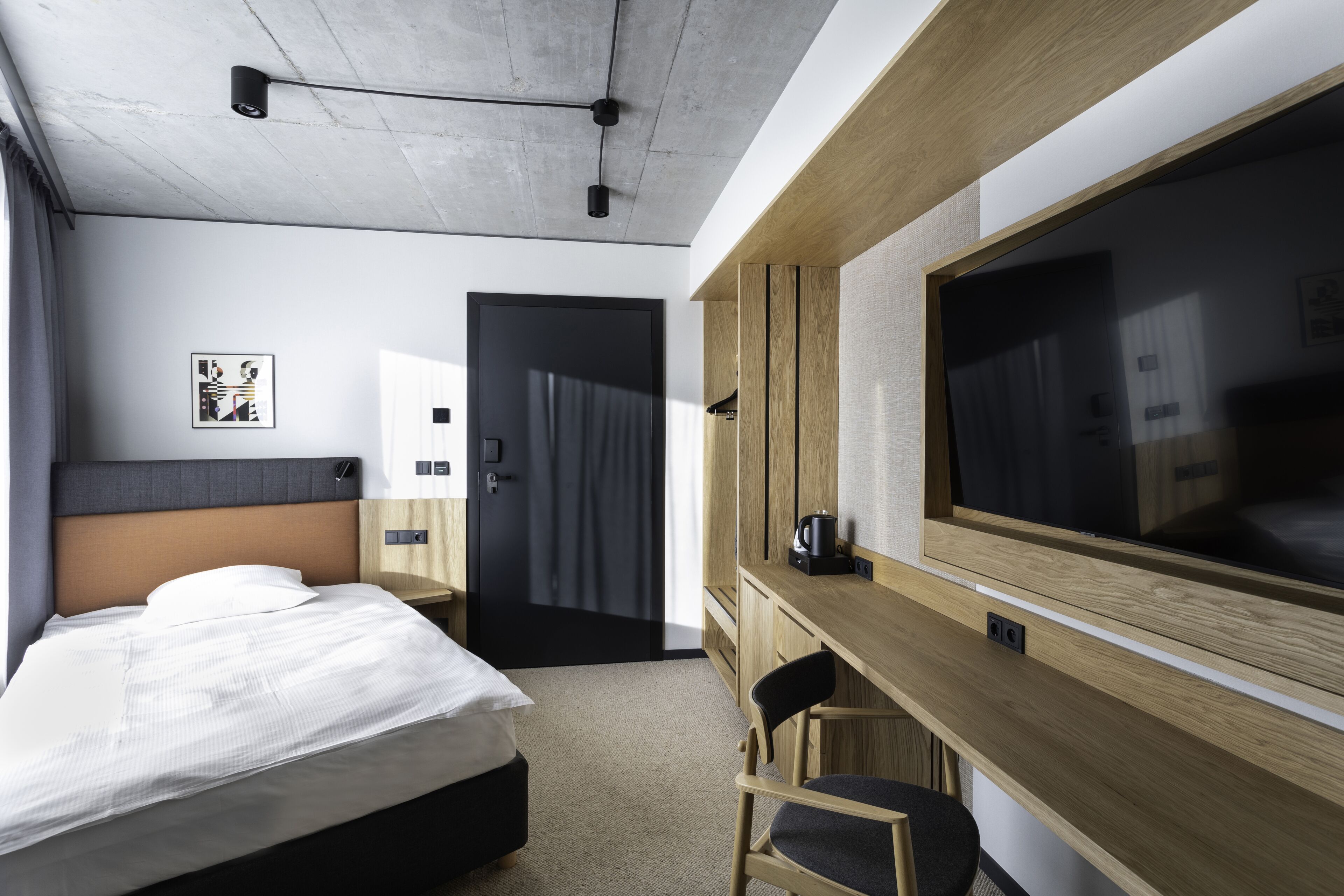 A modern hotel room with a bed, a desk, a chair, a large TV, and a door leading to another room.
